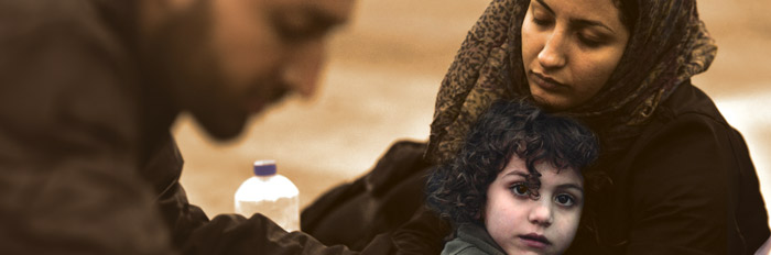 A woman wearing a headscarf sits with a child resting in her lap, while a man is partially visible to the side. The scene conveys a sense of care and compassion in a difficult situation.