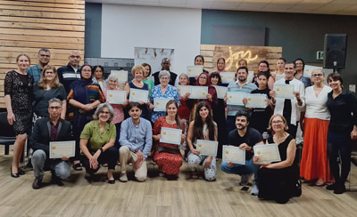 A diverse group of individuals is gathered in a spacious venue, all holding certificates and smiling proudly. The setting includes wooden paneling and decor in soft lighting, indicating a celebratory atmosphere.