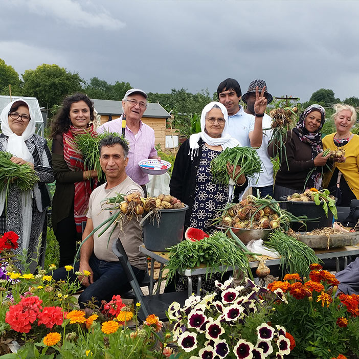 Group of people smiling while holding freshly harvested vegetables in a community garden, surrounded by colorful flowers and a cloudy sky. Community gardening initiatives promote healthy eating and social interaction.