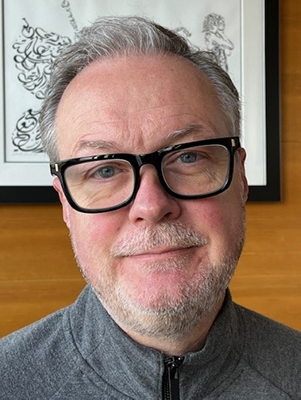 A man with glasses and a slight beard, wearing a gray zip-up sweater, smiling in front of artwork.