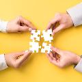 Hands holding four interlocking white puzzle pieces on a vibrant yellow background, symbolizing teamwork and collaboration.