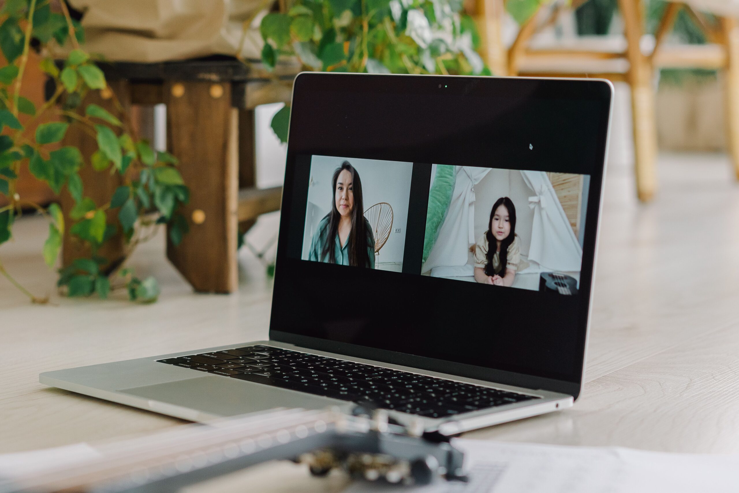 A laptop displays a virtual meeting between a woman and a girl in a home setting with plants.